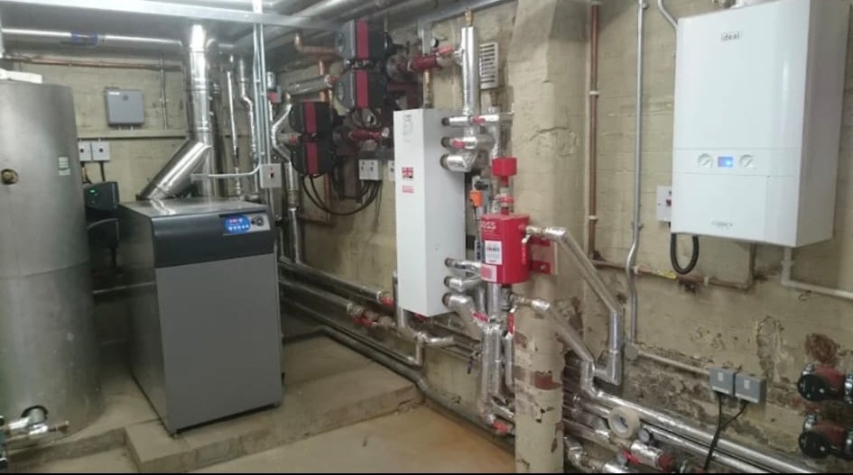 Ideal wall-hung boiler alongside a floor-standing system boiler, hot water cylinder and manifold with structured stainless steel pipework in a commercial plantroom, London.